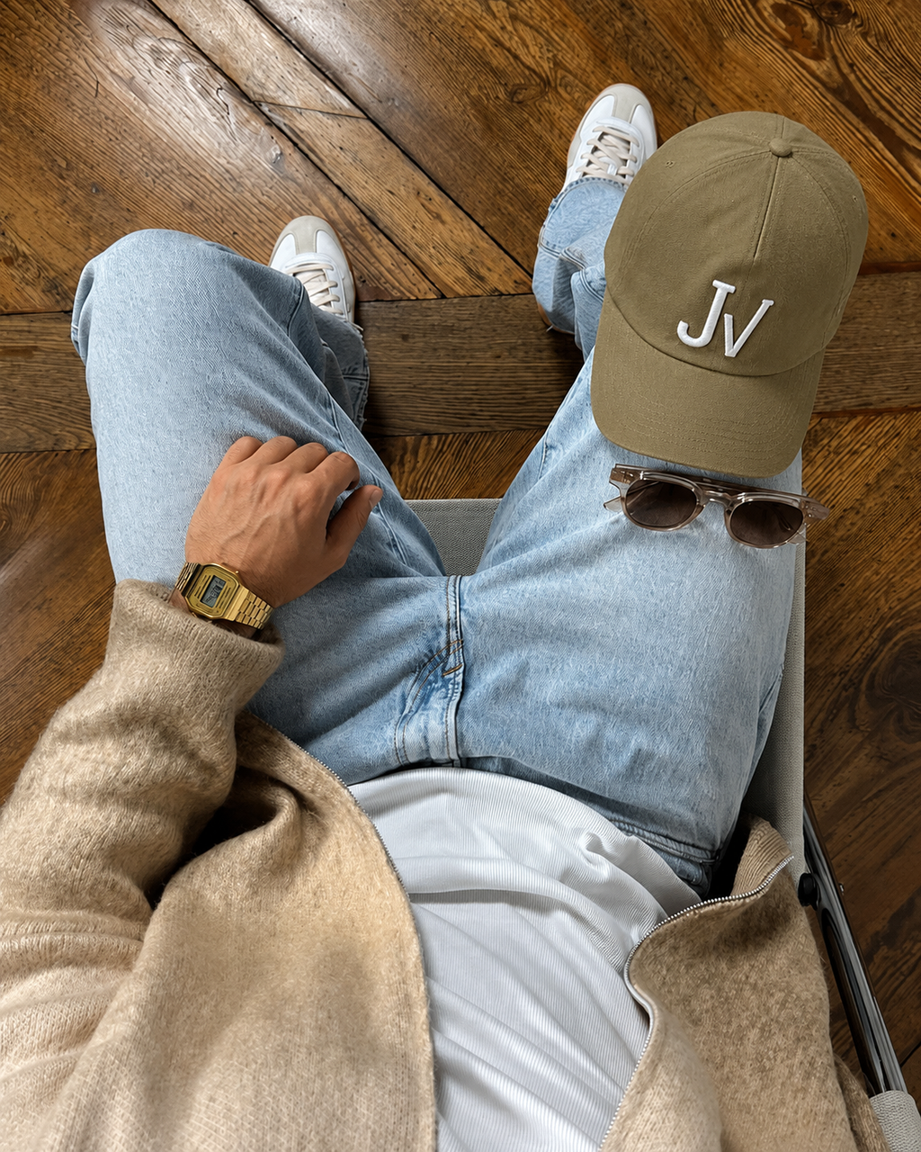 Person sitting on a wooden floor wearing a beige cap with 'JV' logo, light blue jeans, and white sneakers.